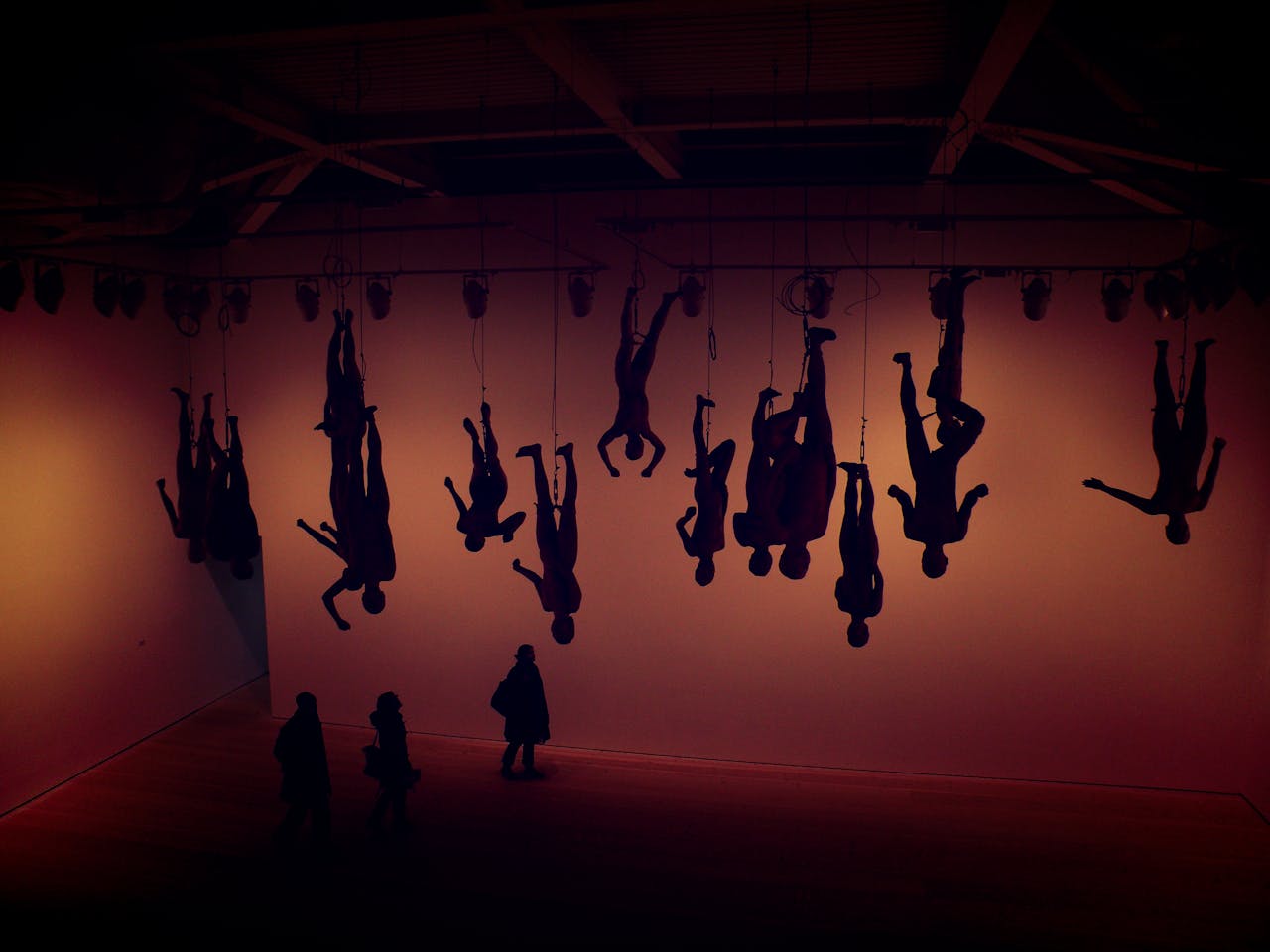 our-services-1 Silhouette of hanging figures in a London gallery with visitors viewing the installation.