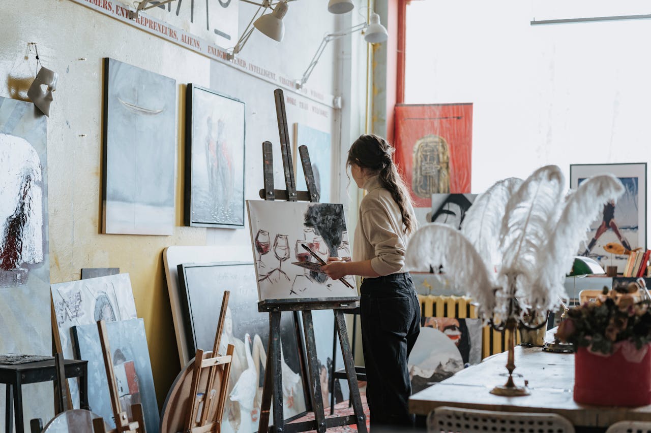 our-services-3 Artist in studio painting with brush on canvas, surrounded by various artworks and decor.